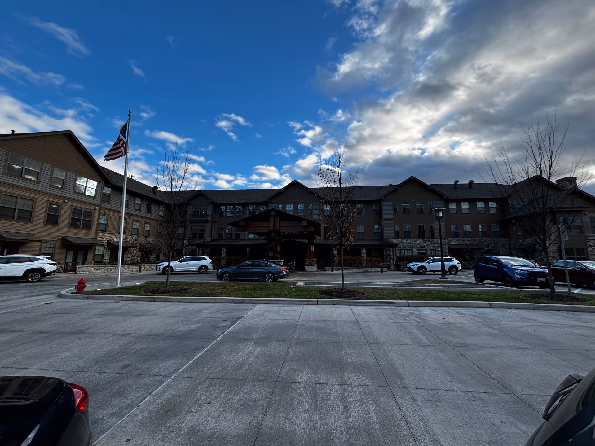 Front exterior view of a large senior living facility building with multiple windows and a covered entrance. Several cars are parked in front, and an American flag is flying on a flagpole near the entrance. The sky is partly cloudy with patches of blue.