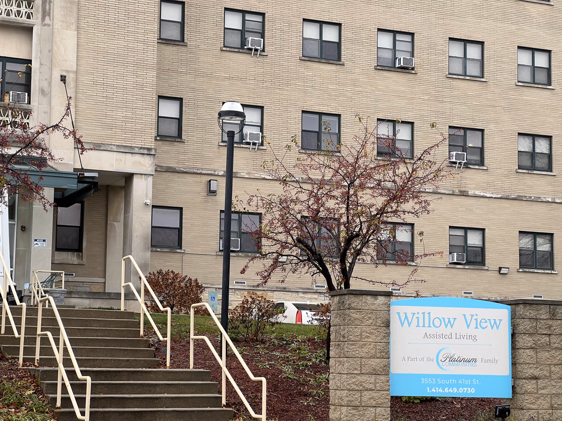 Exterior view of Willow View Assisted Living facility in Milwaukee, showing the building's facade with multiple windows, a staircase with handrails leading up to the entrance, a small tree with red leaves, and a sign displaying the facility's name and contact information.