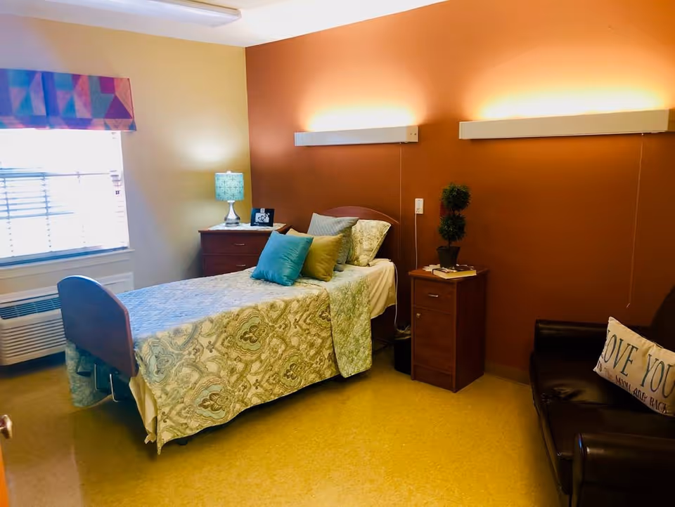 A cozy bedroom in a senior living facility with a single bed covered in a patterned bedspread and several pillows. There is a wooden nightstand with a lamp and a framed photo next to the bed. Another small wooden cabinet with a potted plant and a book is beside the bed. A dark brown leather armchair with a decorative pillow is positioned against the wall. The walls are painted in warm tones, with one wall in a burnt orange color and the others in a light beige. A window with blinds and a colorful valance lets in natural light.