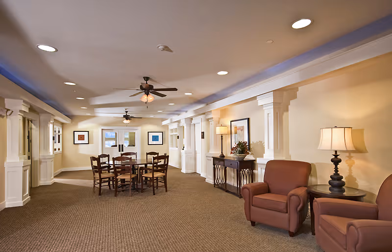 A spacious, well-lit common area with beige walls and carpeted floor. The room features a round wooden table with six chairs in the center, two brown upholstered armchairs with a side table and lamp on the right, and decorative columns along the walls. Ceiling fans with lights are mounted on the ceiling, and framed artwork is hung on the walls.