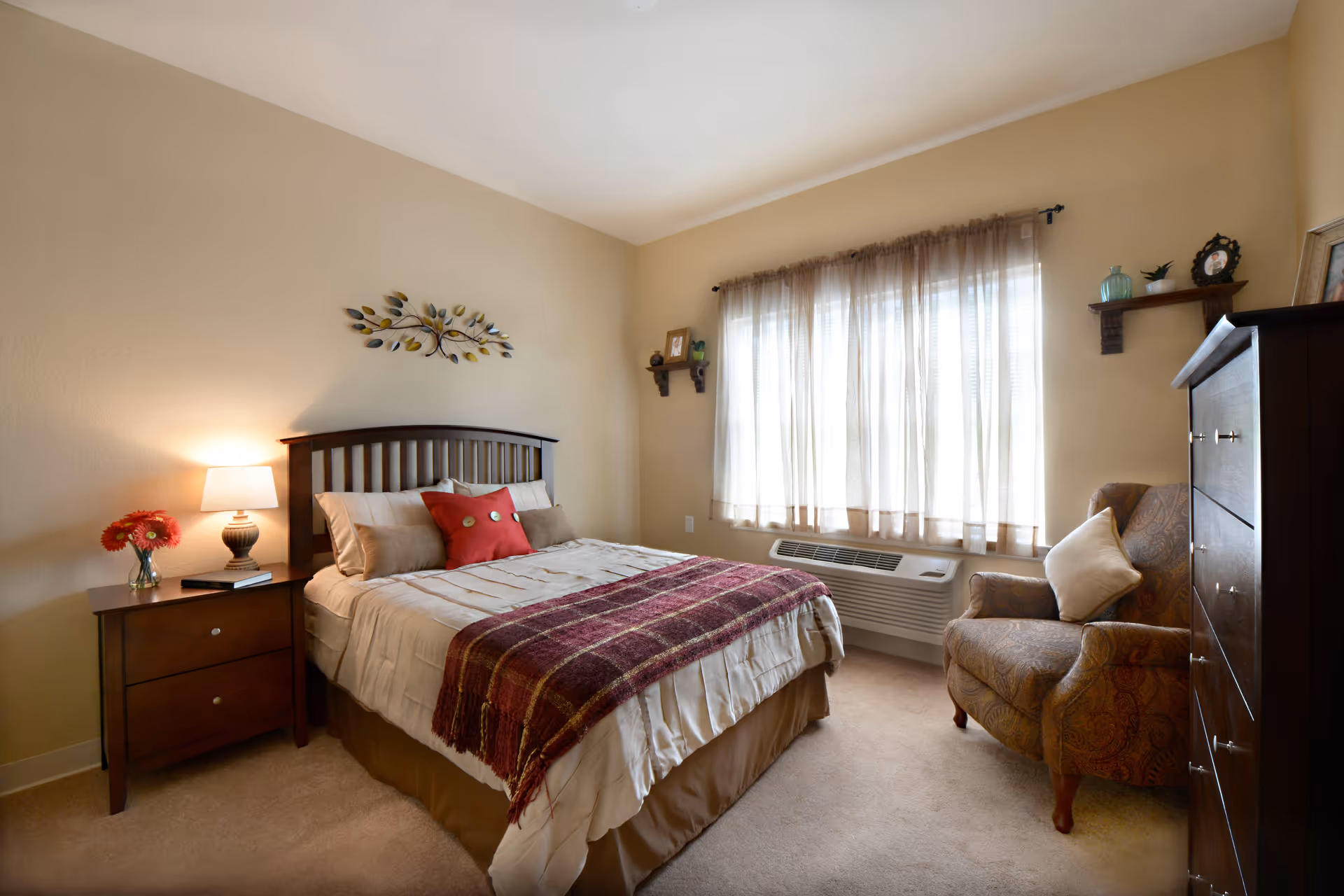 A cozy bedroom with a neatly made bed featuring beige bedding and a red throw blanket. There is a wooden headboard and a nightstand with a lamp and a vase of red flowers. A patterned armchair with a cushion is placed near a window with sheer curtains. A wooden dresser and two small wall shelves with decorative items are also visible.