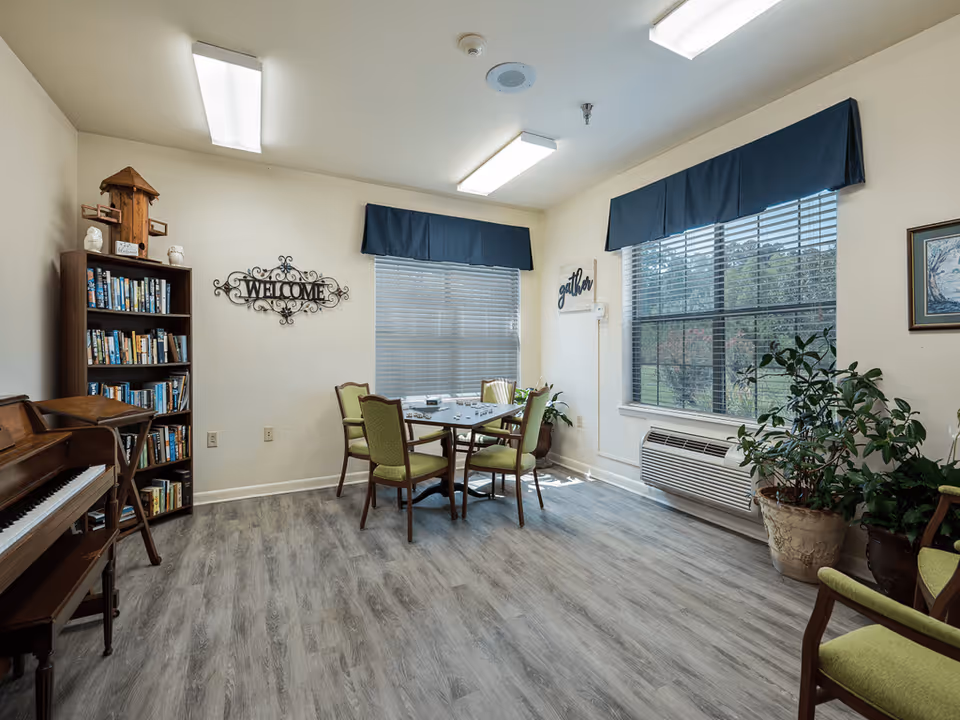 A bright room with light-colored walls and wood-patterned flooring featuring a small table with four green cushioned chairs. There is a bookshelf filled with books, a piano, and several potted plants near large windows with blue valances. Wall decor includes a metal 'WELCOME' sign and a 'gather' sign.