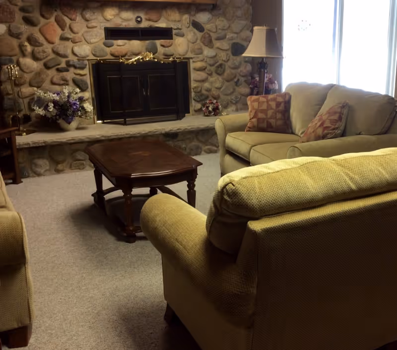 A cozy living room with beige upholstered sofas and a wooden coffee table in the center. The room features a stone fireplace with a black metal screen, decorated with floral arrangements on either side. A tall lamp with a beige shade stands near the window, which lets in natural light.