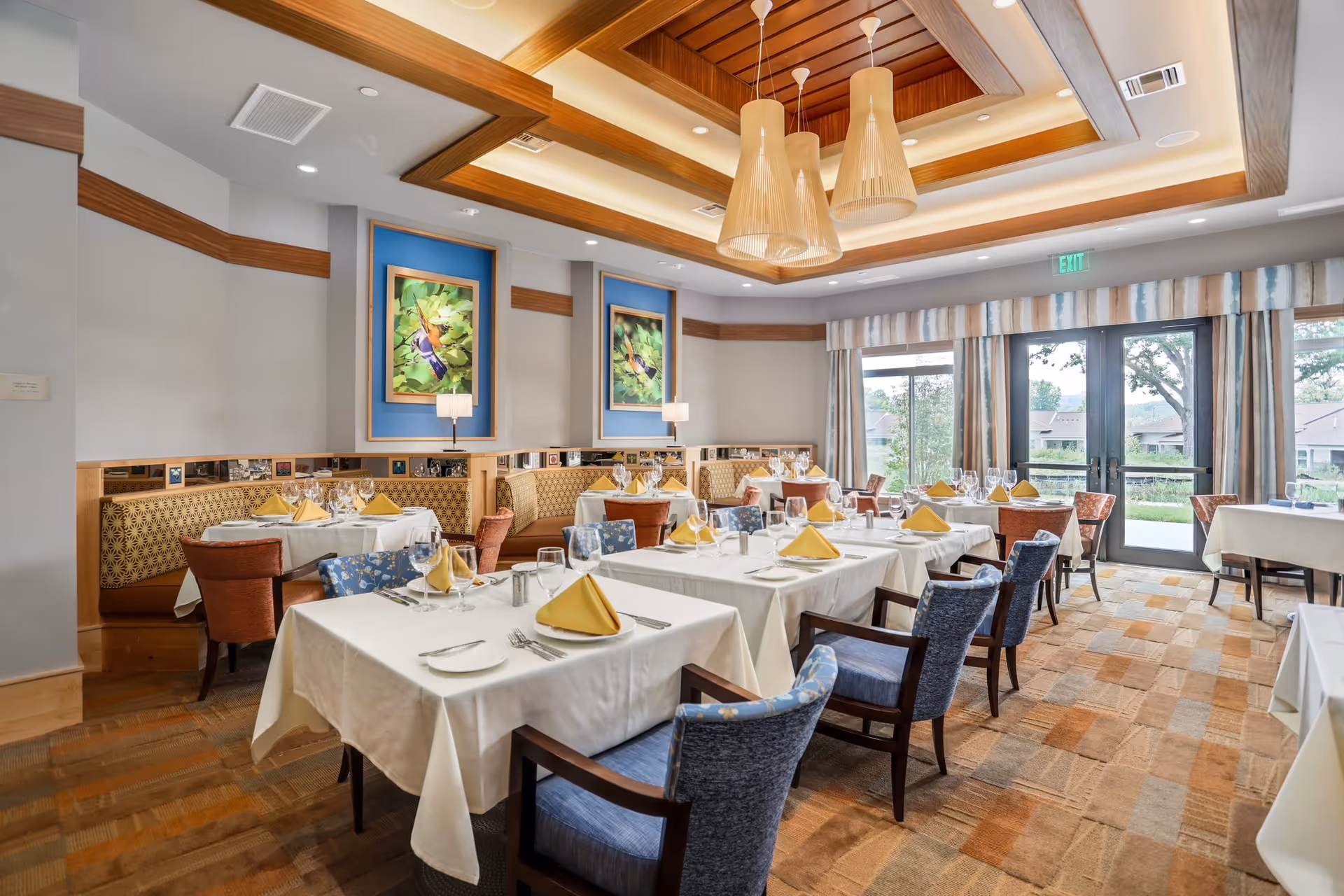 A bright and elegant dining room with tables covered in white tablecloths, set with yellow folded napkins, glassware, and silverware. The room features comfortable upholstered chairs in blue and brown tones, patterned carpet, and large windows with curtains allowing natural light to enter. The ceiling has wooden beams and modern pendant lights, and colorful framed artwork is displayed on the walls.
