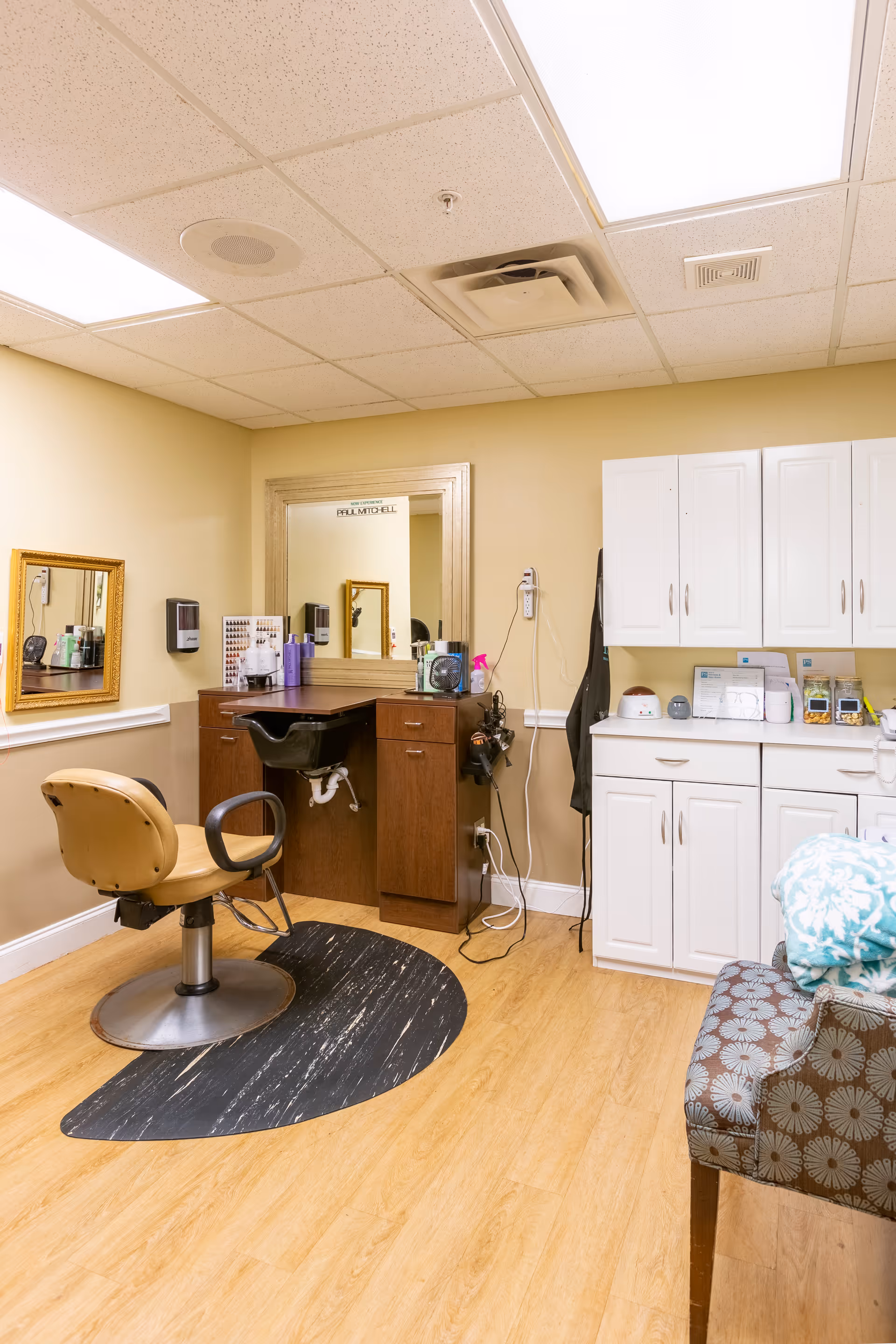 A small salon area inside a senior living facility with a salon chair in front of a wooden vanity with a mirror. The vanity has hair care products and a hair washing sink. To the right, there are white cabinets and a countertop with various small appliances and items. A patterned chair with a folded blanket is partially visible in the foreground. The room has beige walls and a light wood floor.