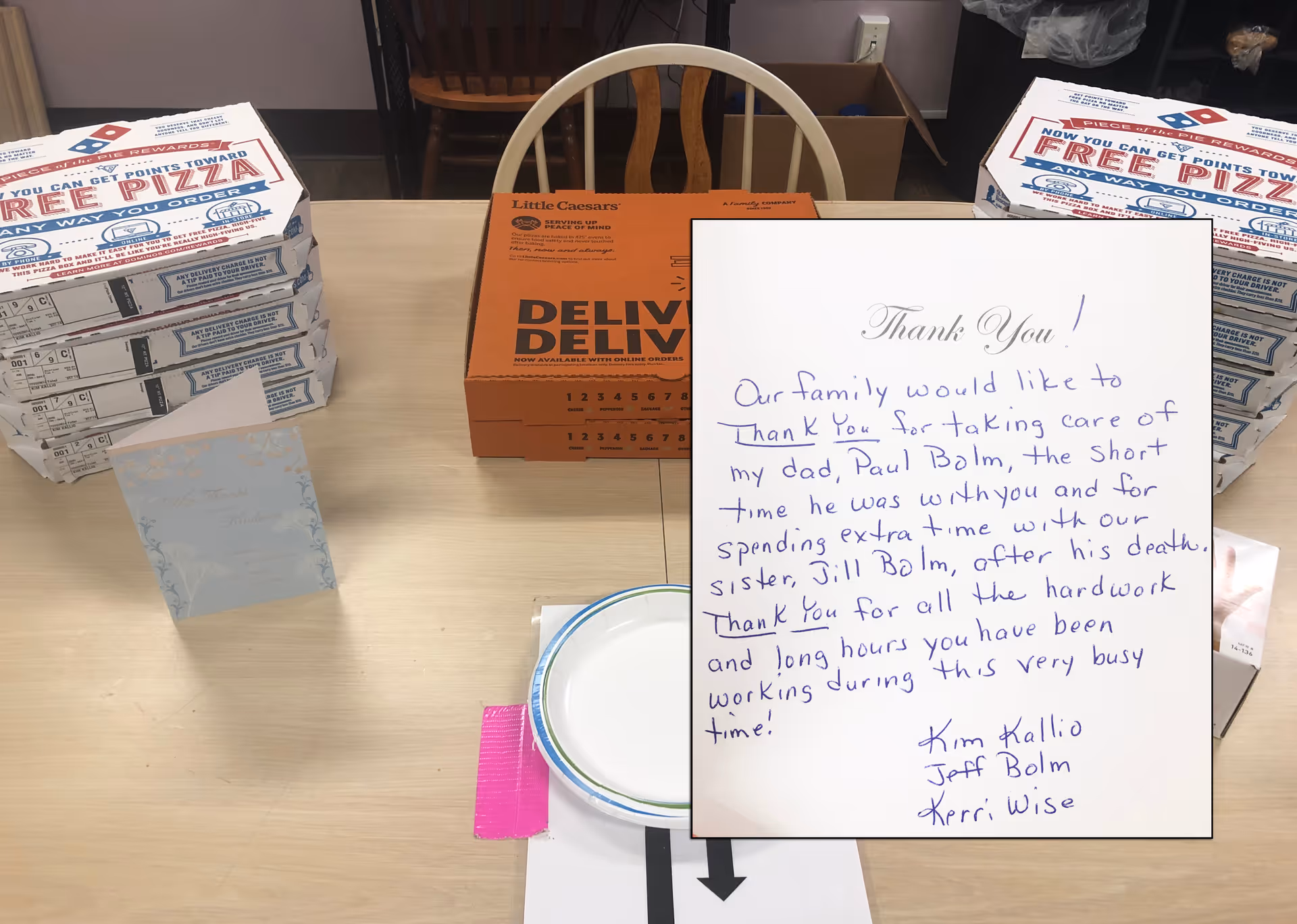 A table in a dining area with stacked pizza boxes, a Little Caesars box, a thank-you card, and paper plates.