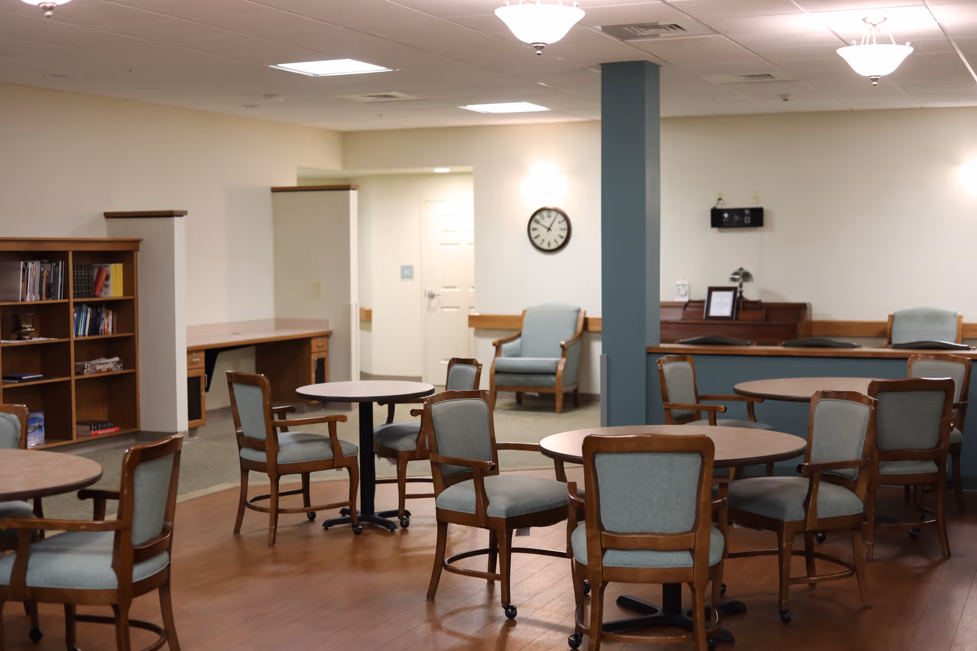 A well-lit common area with several round tables and wooden chairs with light blue cushions. There is a bookshelf with books on the left side, a clock on the wall, and a piano against the far wall. The floor is wooden, and the walls are painted light beige with a teal column in the middle of the room.