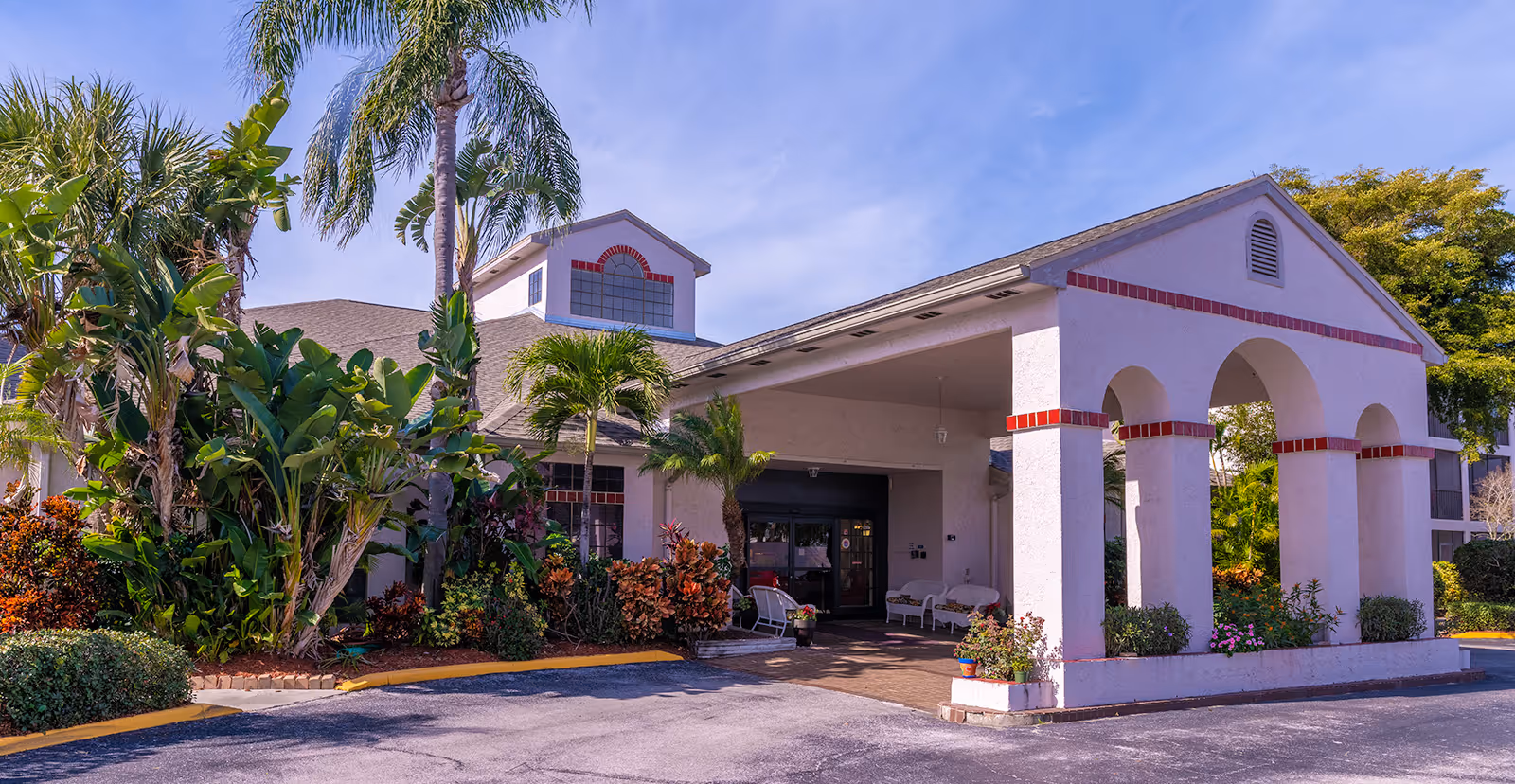 Front entrance of a senior living building with a covered porte-cochère, arched columns, and tropical landscaping.