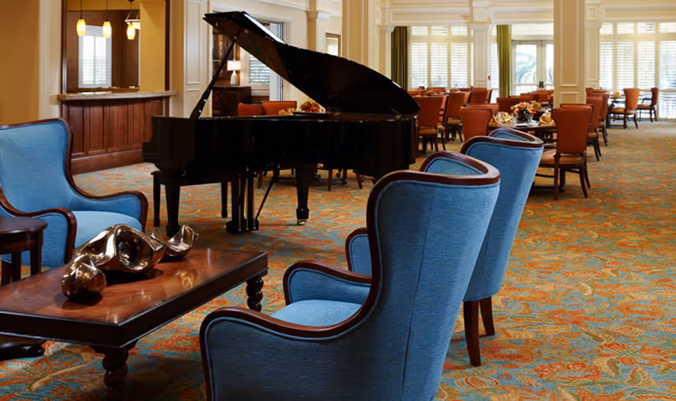 A spacious interior room featuring a black grand piano in the center, surrounded by blue upholstered armchairs and a wooden coffee table with decorative items. In the background, there are multiple dining tables with orange chairs, set for a meal, and large windows with blinds letting in natural light.