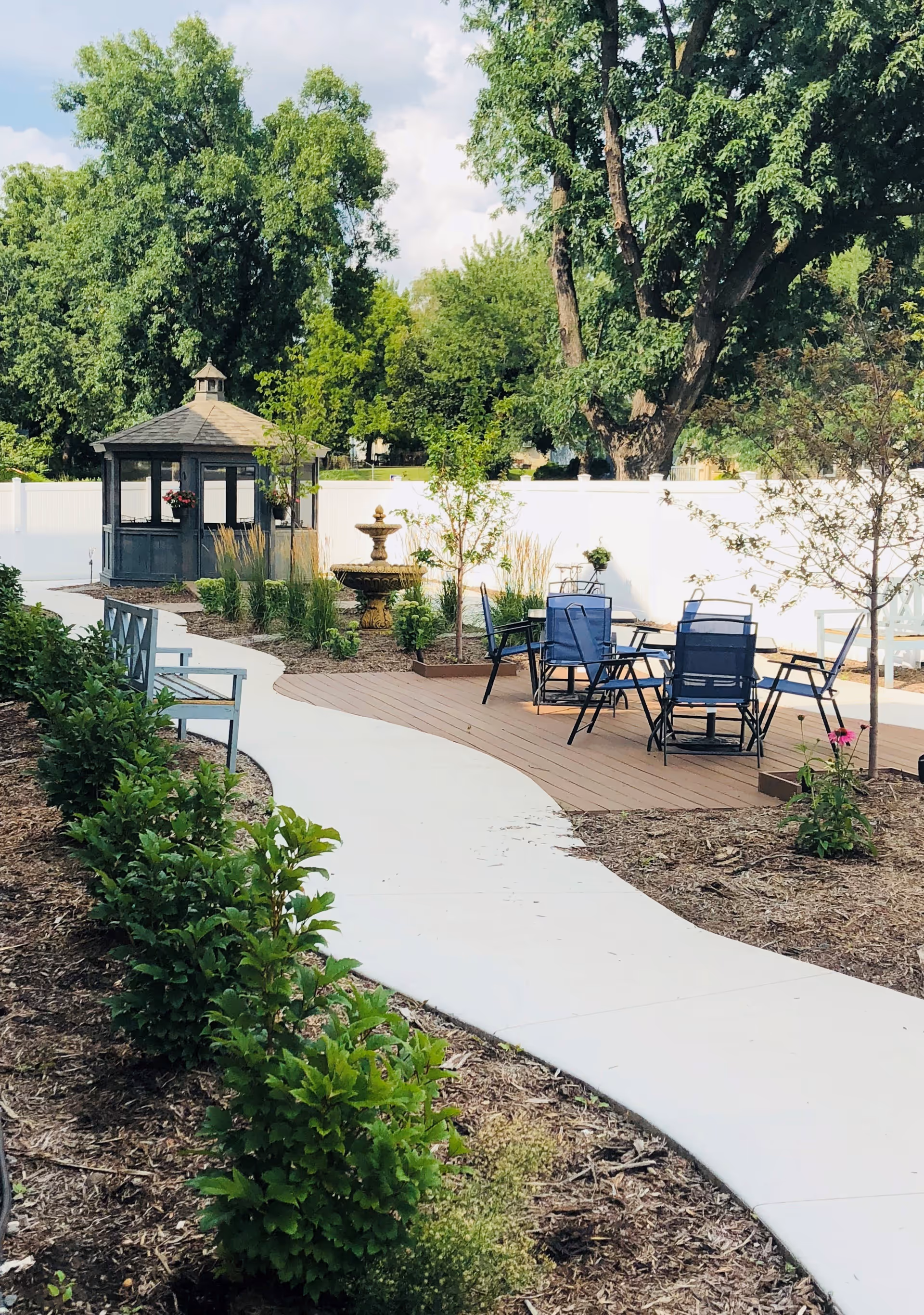 Outdoor garden area with a winding concrete pathway, green bushes along the path, a wooden deck with blue chairs and tables, a small gazebo, and a decorative fountain, surrounded by trees and a white fence.