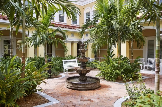 Sunlit courtyard with a central tiered fountain surrounded by palm trees, benches, and yellow building facades.