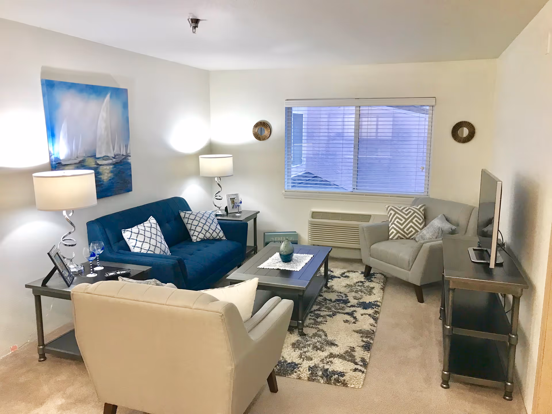 Neatly furnished living room with a blue sofa, two armchairs, coffee table, TV and lamps under a window.