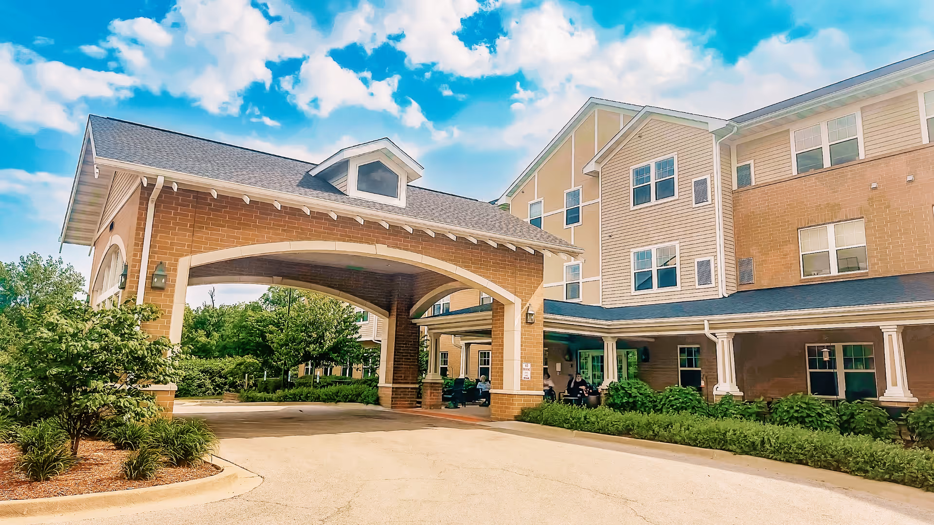 Front entrance of a multi-story senior living building with a covered drive-up portico, columns, and surrounding landscaping.