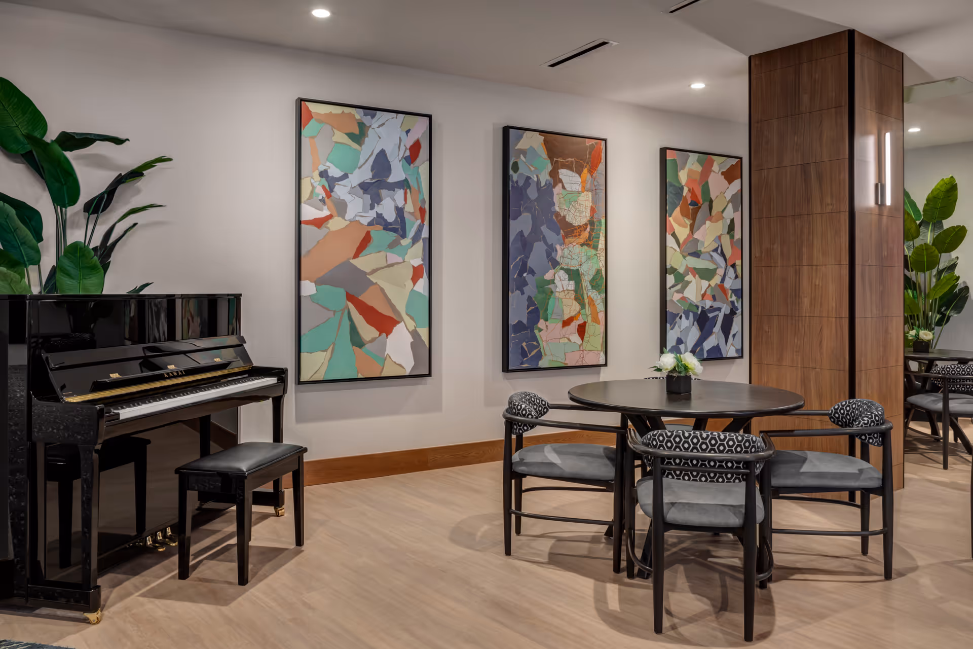 A modern interior room featuring a black upright piano with a matching bench on the left, a round black table with four patterned cushioned chairs around it, and three colorful abstract paintings hanging on the wall. There are large green plants on both sides of the room and a wooden column with a vertical light fixture.