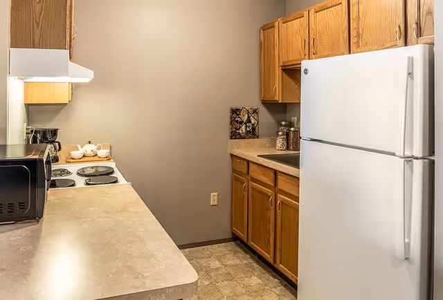 A compact kitchen with wooden cabinets, a white refrigerator, a microwave, a coffee maker, and a stove with four burners. The countertops are beige, and the floor has a tiled pattern. The walls are painted a neutral beige color.