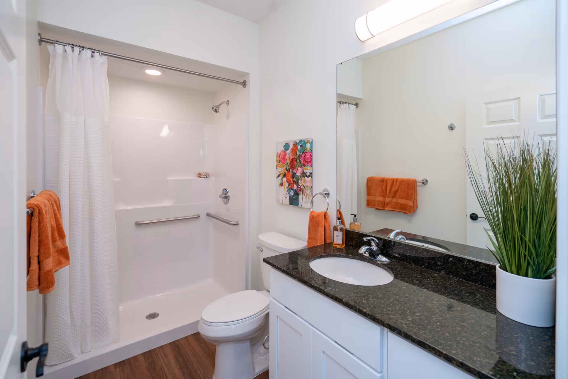 A clean and modern bathroom featuring a white shower with a curtain, a toilet, a black granite countertop with a sink, a large mirror, orange towels, a colorful floral painting on the wall, and a green potted plant on the counter.