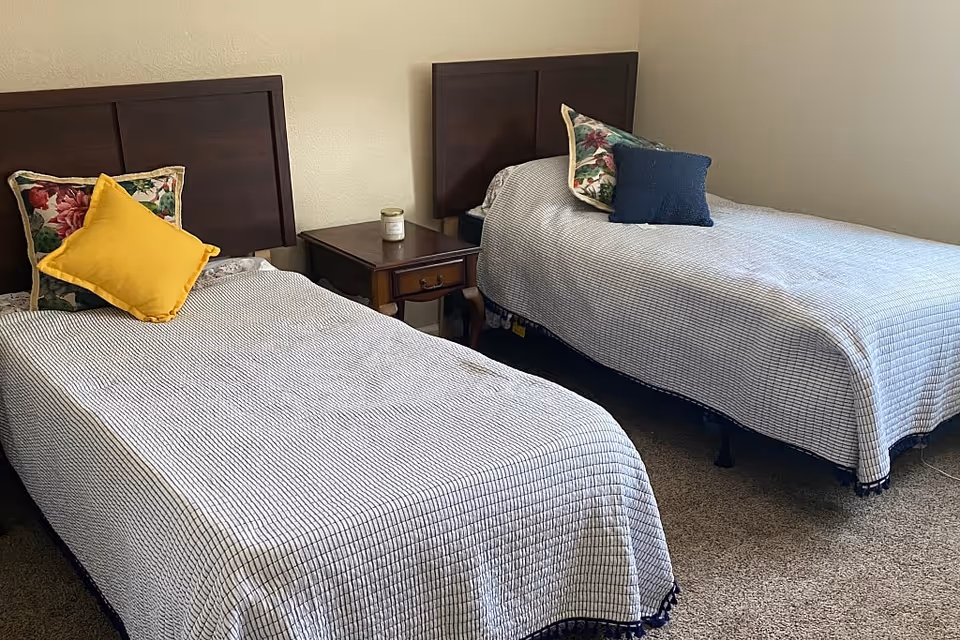 A bedroom with two twin beds, each with a dark wooden headboard. Both beds have white and black checkered bedspreads and decorative pillows, one with a yellow pillow and floral pillow, the other with a blue pillow and floral pillow. A wooden nightstand with a candle is placed between the beds. The room has beige walls and carpeted flooring.