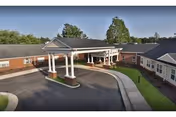 Exterior view of a single-story healthcare facility building with a covered entrance supported by white columns. The building has a brick facade and is surrounded by a paved driveway and green landscaping with trees in the background.