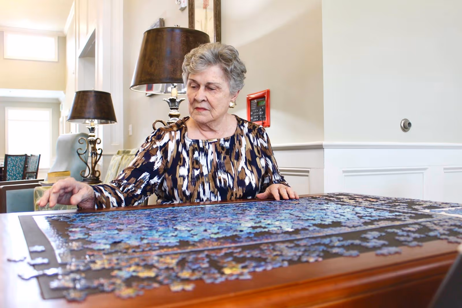An elderly woman sitting at a wooden table working on a large jigsaw puzzle in a well-lit room with lamps and chairs in the background.