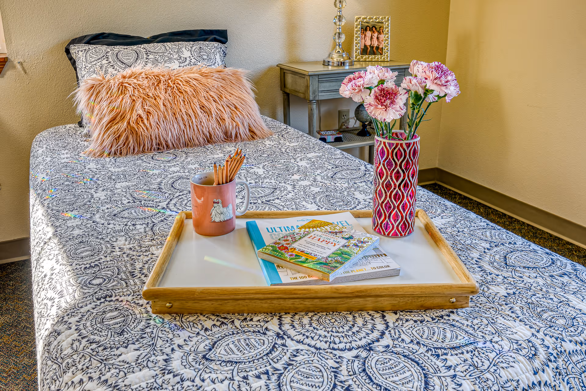 A neatly made single bed with a patterned bedspread and a fluffy pink pillow. On the bed is a wooden tray holding a pink mug with colored pencils, a vase with pink and white flowers, and a few books. Next to the bed is a small gray nightstand with a silver lamp and a framed photo.