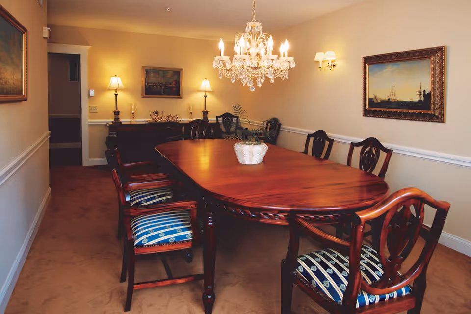 Formal dining room with a long polished wooden table, upholstered chairs, a crystal chandelier, and framed paintings on the walls.