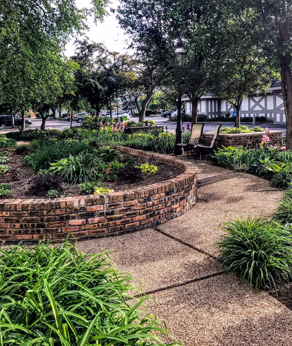 A landscaped outdoor garden area with a curved brick planter filled with various green plants and flowers. There is a paved walkway leading to a seating area with two chairs and a lamp post. Trees provide shade and a building with a Tudor-style exterior is visible in the background along with parked cars.