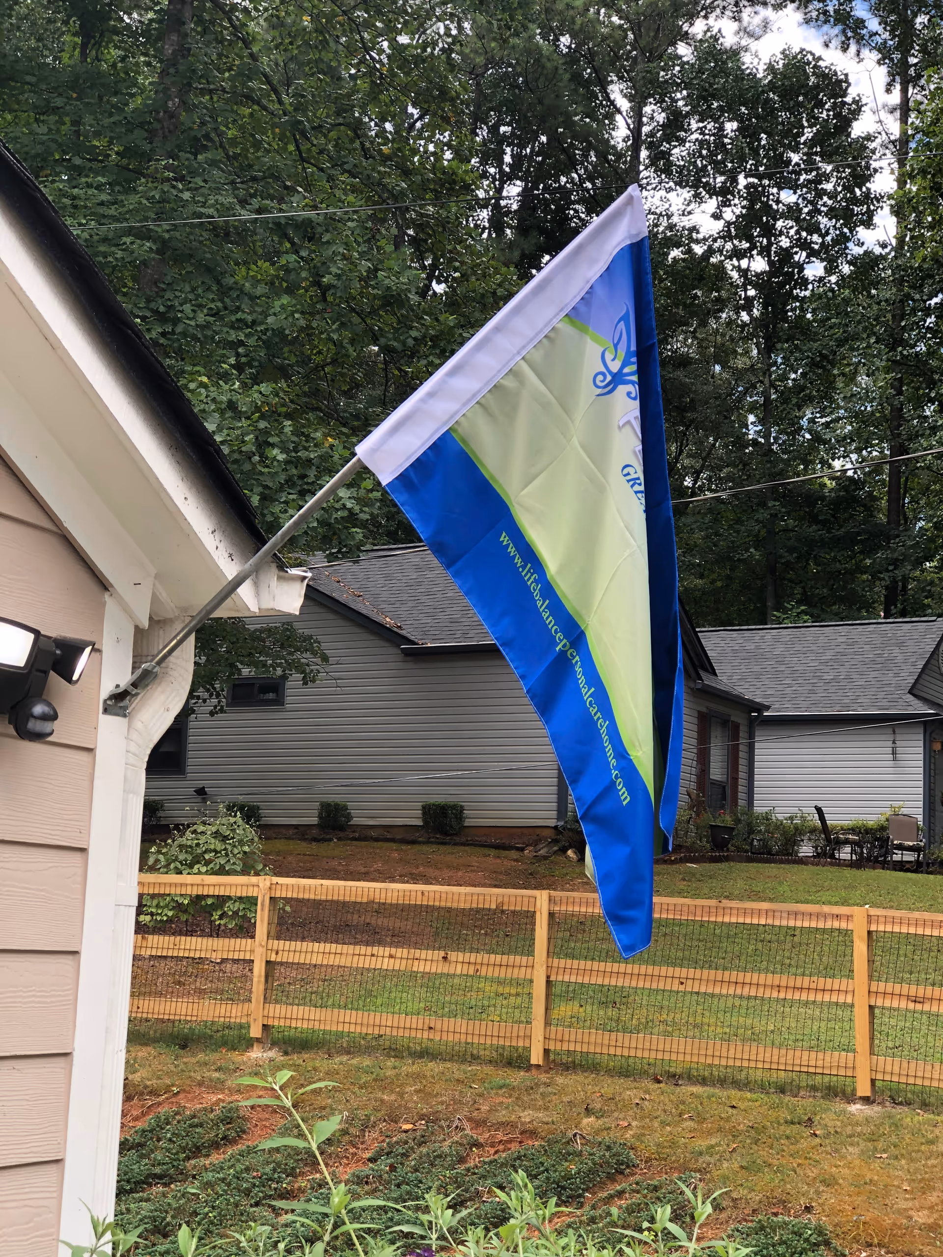 Blue and green flag mounted on the exterior of a house over a fenced yard with neighboring houses and trees in the background.
