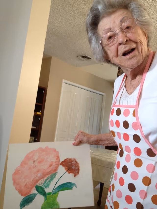 An elderly woman wearing glasses and a white shirt with a pink and brown polka dot apron is holding a painting of two flowers in a green vase. She is indoors in a room with beige walls, a table with a tablecloth, and a white closet door in the background.