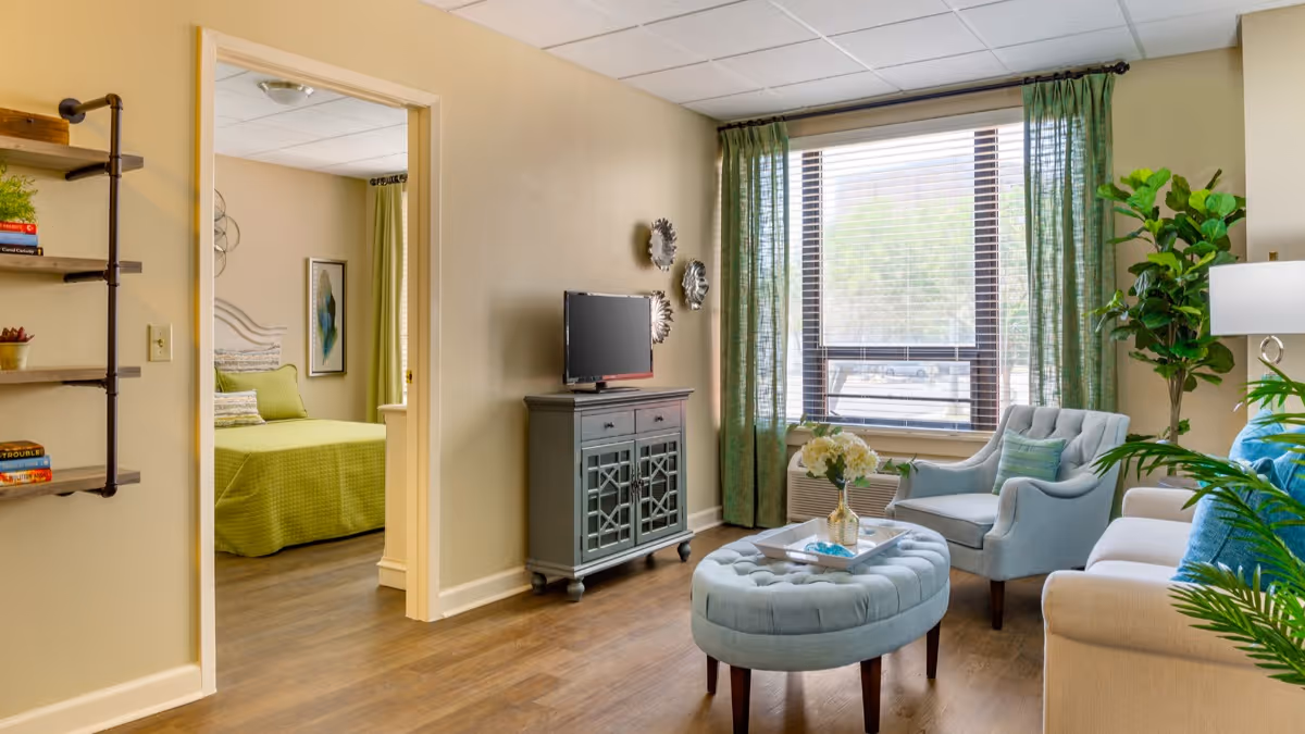 A bright living room with a large window covered by green curtains, a light blue armchair, a beige sofa with blue cushions, a round tufted ottoman with a tray and flowers, a TV on a gray cabinet, and a view into a bedroom with a green bedspread and wall art.