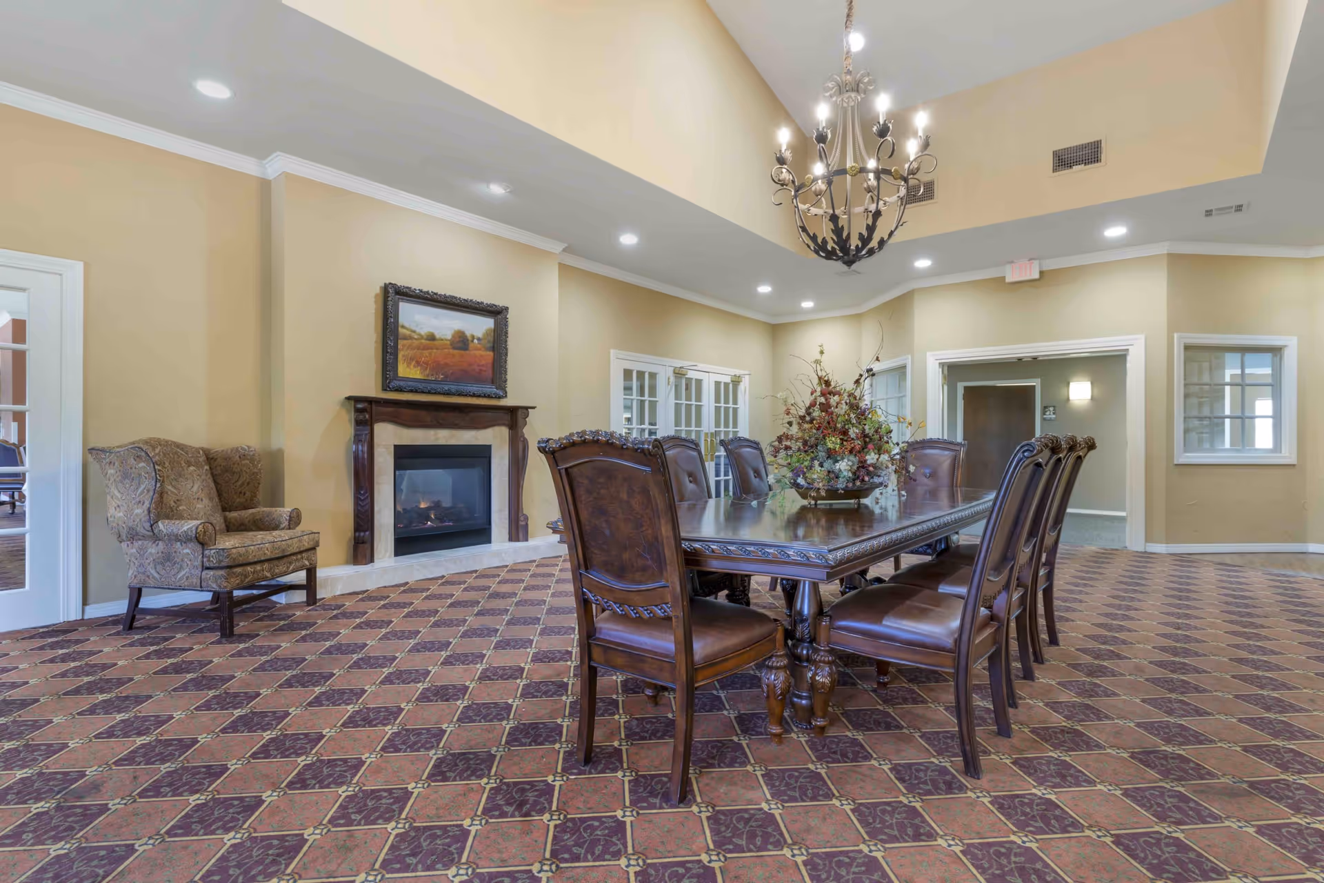 A spacious, elegant room with a large wooden dining table surrounded by eight matching chairs. The room features a patterned carpet, a fireplace with a framed landscape painting above it, a comfortable upholstered armchair, and a decorative chandelier hanging from a high ceiling. The walls are painted beige, and there are multiple doors and windows with white trim.