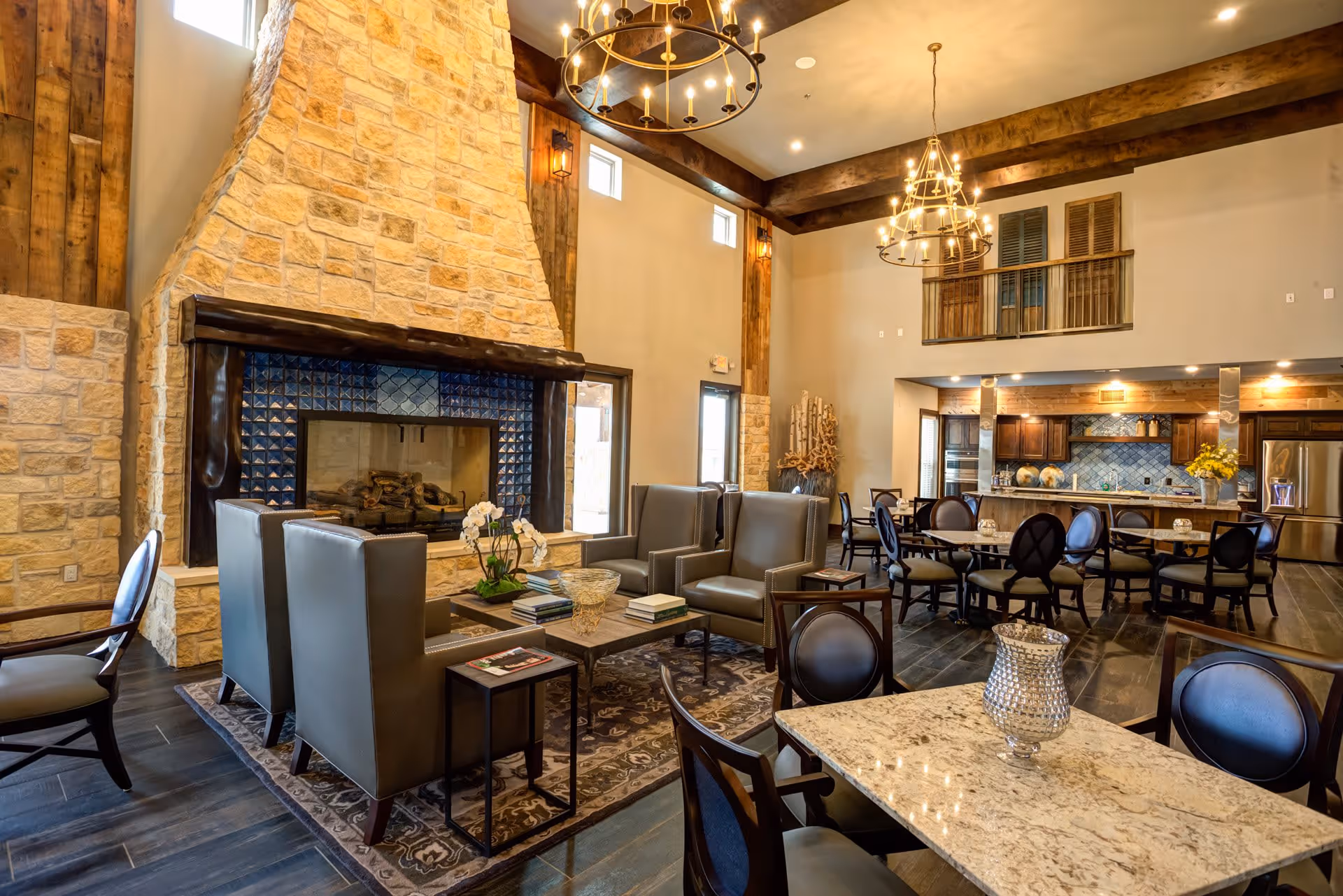 Spacious senior living common area with a large stone fireplace, leather armchairs arranged around a coffee table, multiple dining tables and chairs, wooden ceiling beams, and warm lighting from chandeliers.