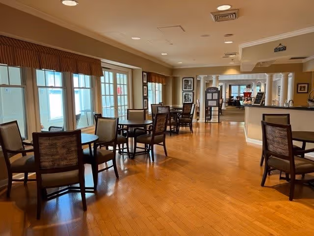 A spacious dining area with multiple round tables and chairs arranged neatly on a wooden floor. Large windows with valances allow natural light to fill the room. The area extends into another room with more seating and a counter or reception area on the right side.