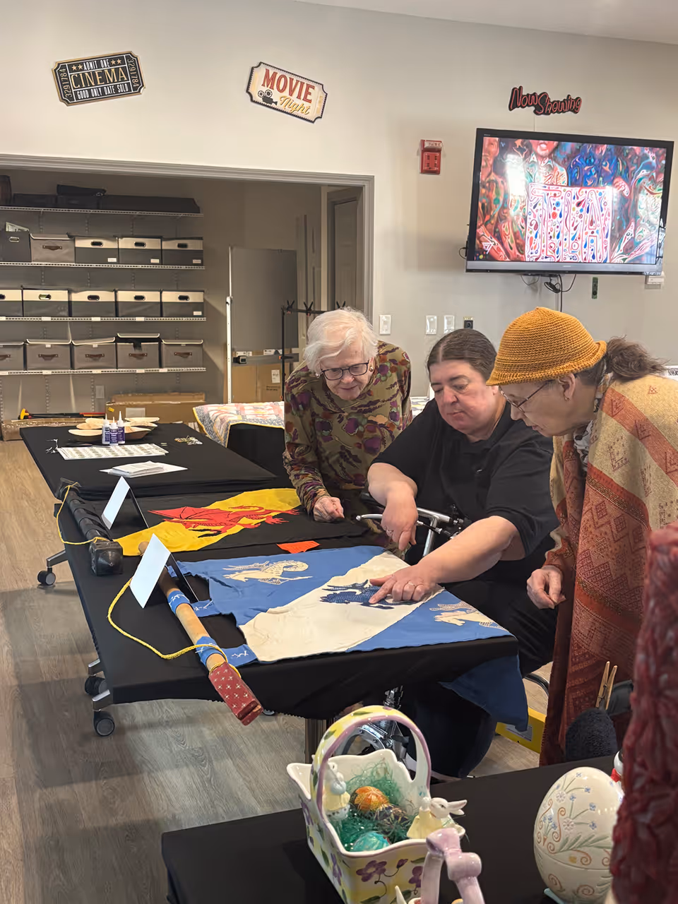 Three elderly women gathered around a table looking at colorful fabric banners or flags laid out on the table in a room with shelves and a TV screen displaying artwork. The room has signs on the wall reading 'Cinema', 'Movie Night', and 'Now Showing'.
