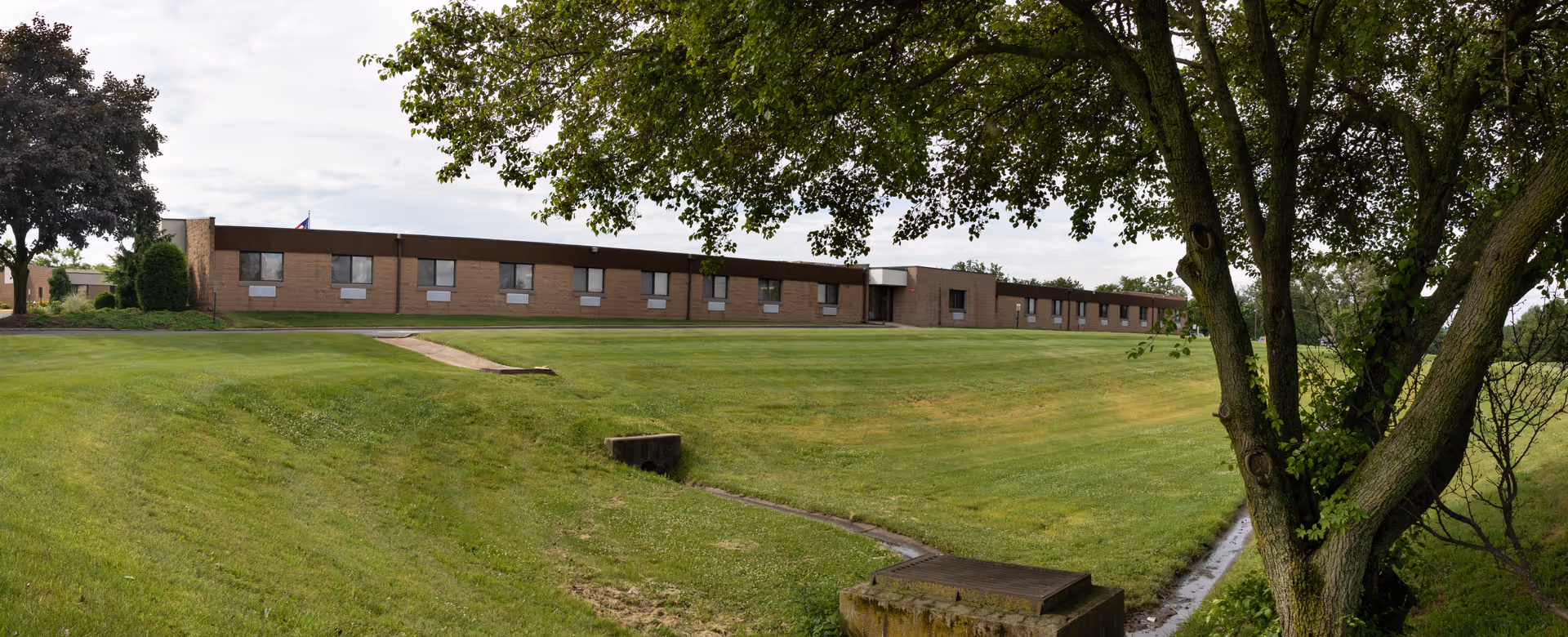 A single-story brick building with multiple windows, set behind a large grassy lawn with a concrete pathway leading towards the entrance. A large tree with green leaves is in the foreground on the right side, partially framing the building. The sky is partly cloudy.