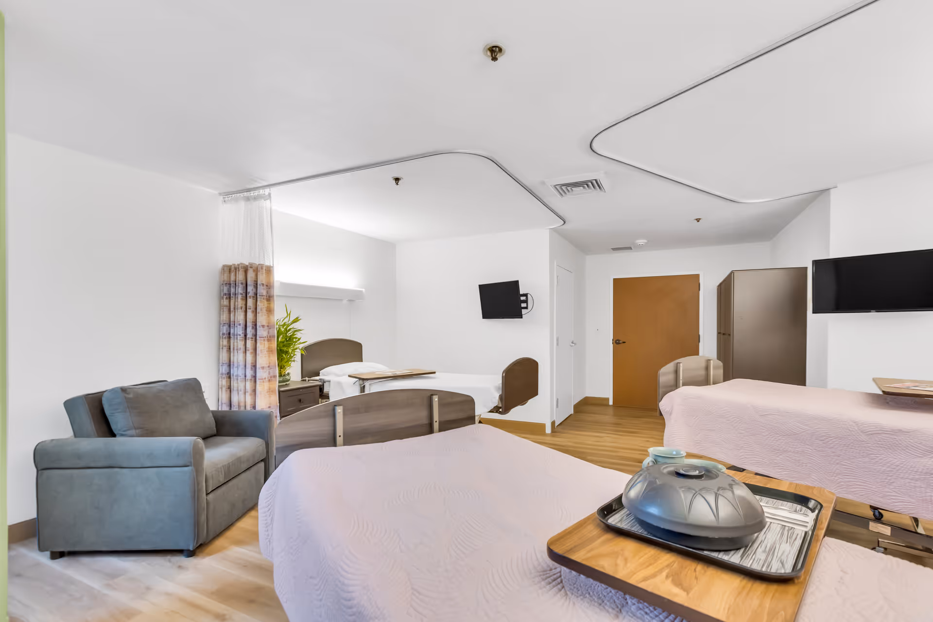 A bright and clean skilled nursing room with two hospital beds covered in light pink bedspreads, a gray armchair, a small wooden nightstand with a plant, two wall-mounted flat-screen TVs, a wooden door, and a cabinet. The room has white walls, light wood flooring, and ceiling-mounted curtain tracks.