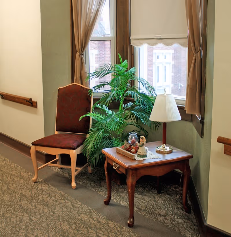 A hallway seating area with a chair, wooden side table, lamp, and potted plant in front of windows.