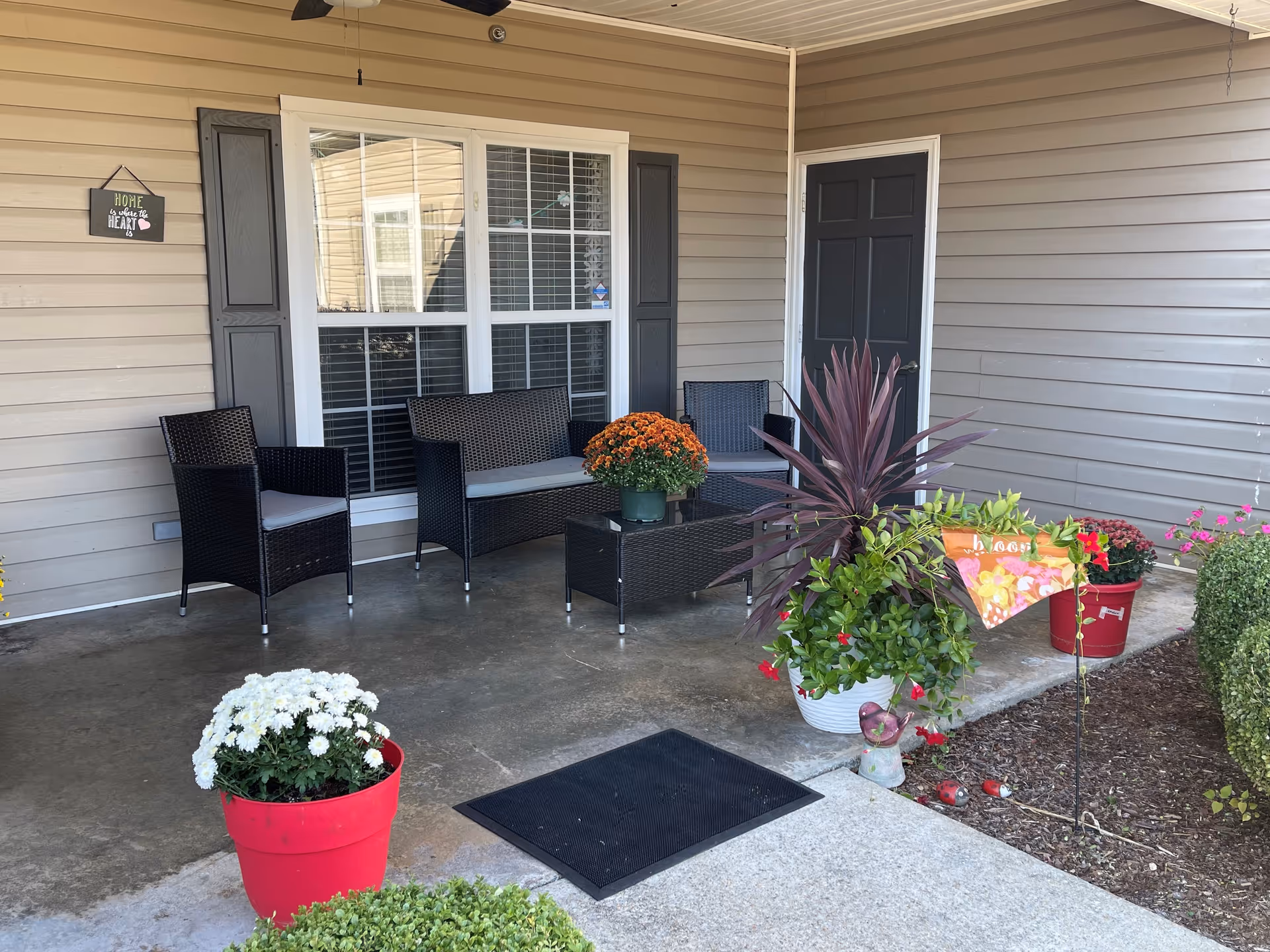A welcoming porch at Gardens of Pelham featuring black wicker chairs, a small table, colorful potted plants, and a decorative sign that reads 'Home is where the heart is'.