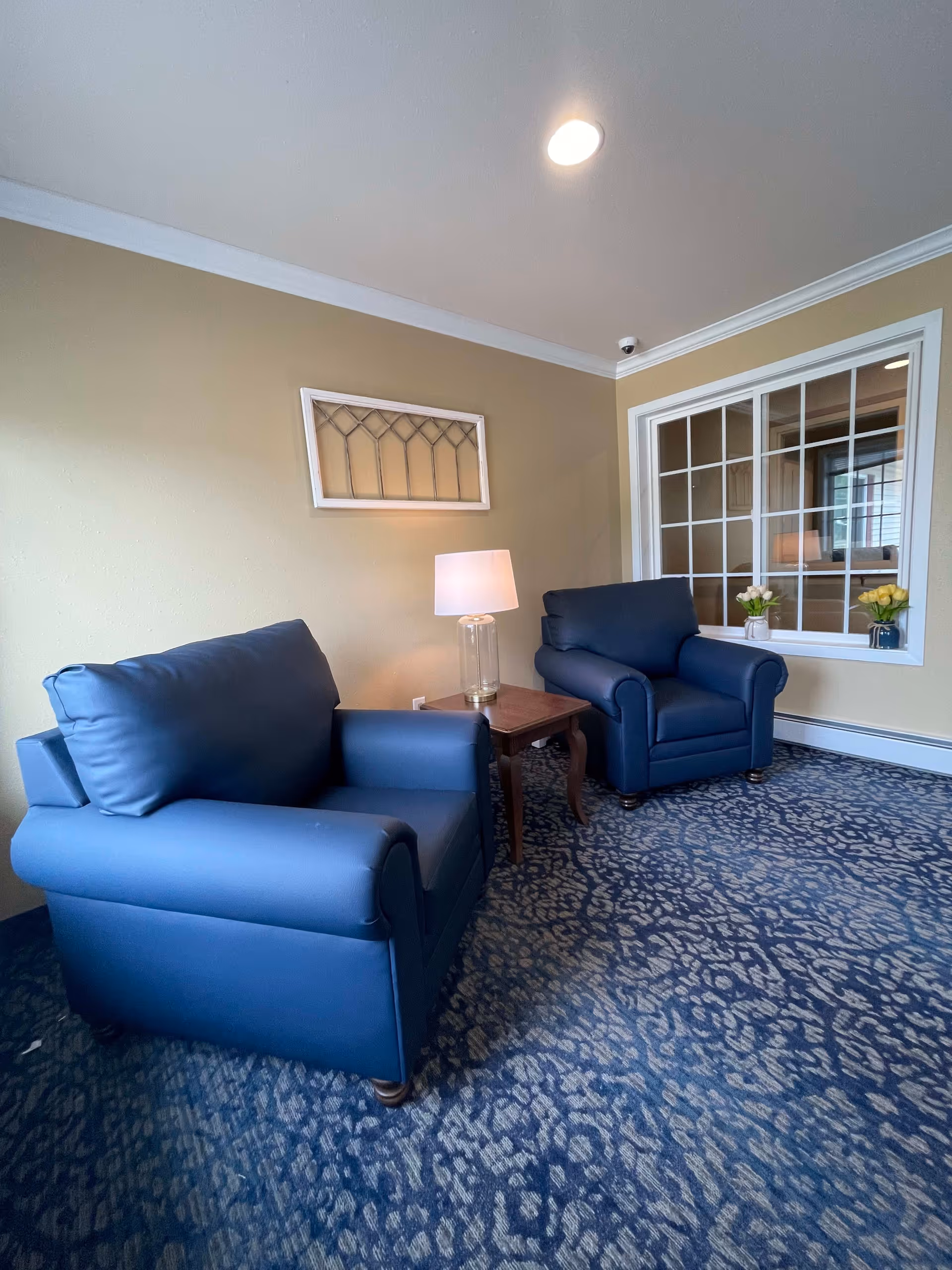 A cozy seating area with two blue armchairs separated by a wooden side table with a lamp on it. The room has beige walls, a patterned blue carpet, and a window with white trim showing another room with flowers on the windowsill.