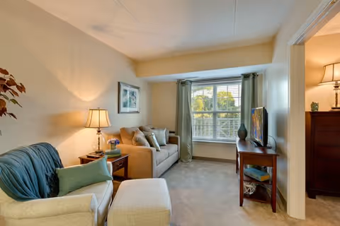 A cozy living room with a beige sofa, a cushioned armchair with a matching ottoman, a wooden side table with a lamp and flowers, a wooden TV stand with a flat-screen TV, and a large window with green curtains letting in natural light.
