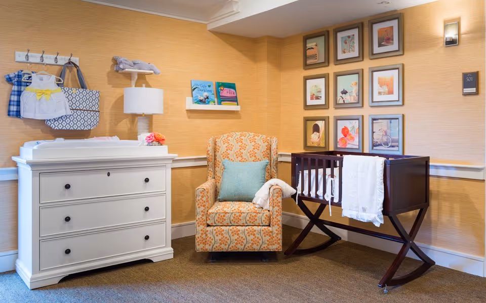 Cozy nursery corner with a white changing-dresser, patterned armchair, wooden cradle, and framed artwork on beige walls.