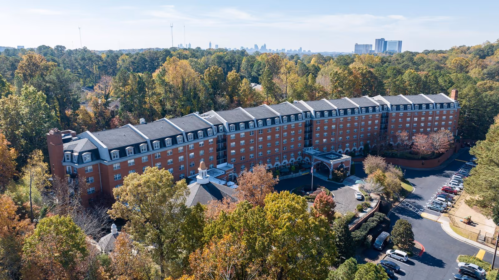 Aerial view of King's Bridge Retirement Community, a large multi-story brick building surrounded by trees with a parking lot visible on the right side. The building has a classic architectural style with multiple windows and a covered entrance. The surrounding area is lush with greenery and the city skyline can be seen faintly in the background.