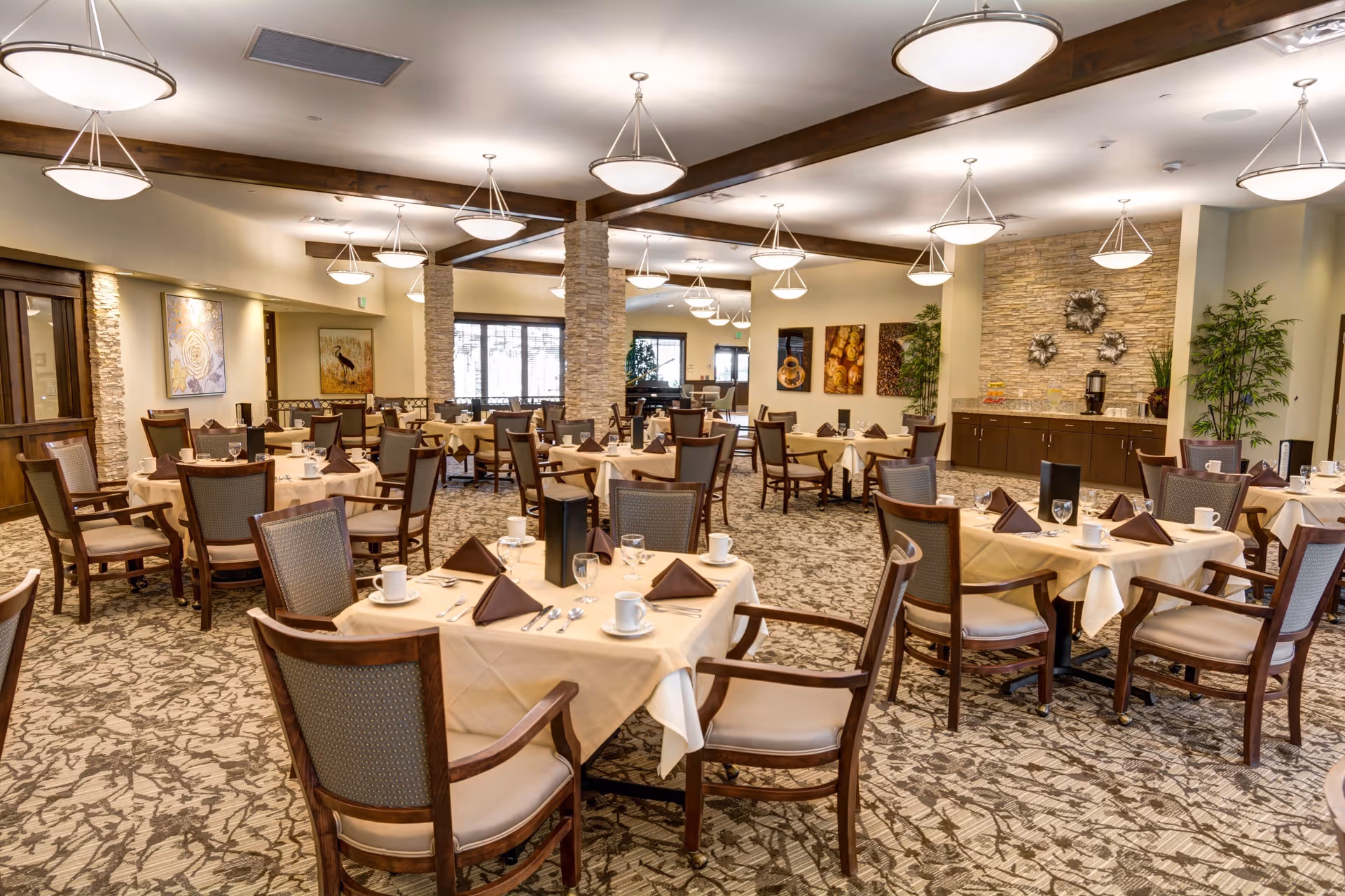 A spacious dining room with multiple round tables covered with beige tablecloths, each set with brown napkins, white cups, glasses, and silverware. The room features patterned carpet, stone pillars, warm lighting fixtures hanging from the ceiling, and decorative artwork on the walls. There is a buffet area with a coffee dispenser and plants in the background.