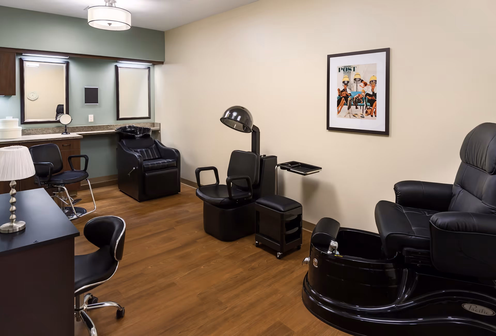 Interior view of a salon room in a senior living facility with black salon chairs, a hair washing station, a hair dryer chair, a pedicure chair with a foot bath, wooden flooring, a framed picture on the wall, and a desk with a lamp and chair.