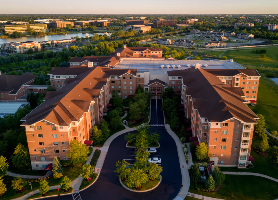 Aerial view of a U-shaped brick senior living complex surrounded by trees, lawns and nearby water with a central driveway and parking.