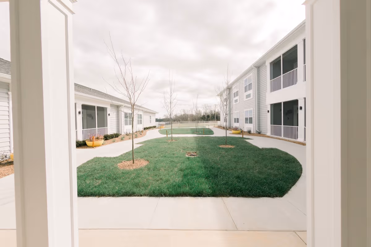View of an outdoor courtyard area at Bluegrass Senior Living, featuring a green lawn with young trees planted in mulch beds, surrounded by a concrete walkway and two-story buildings with balconies and windows.