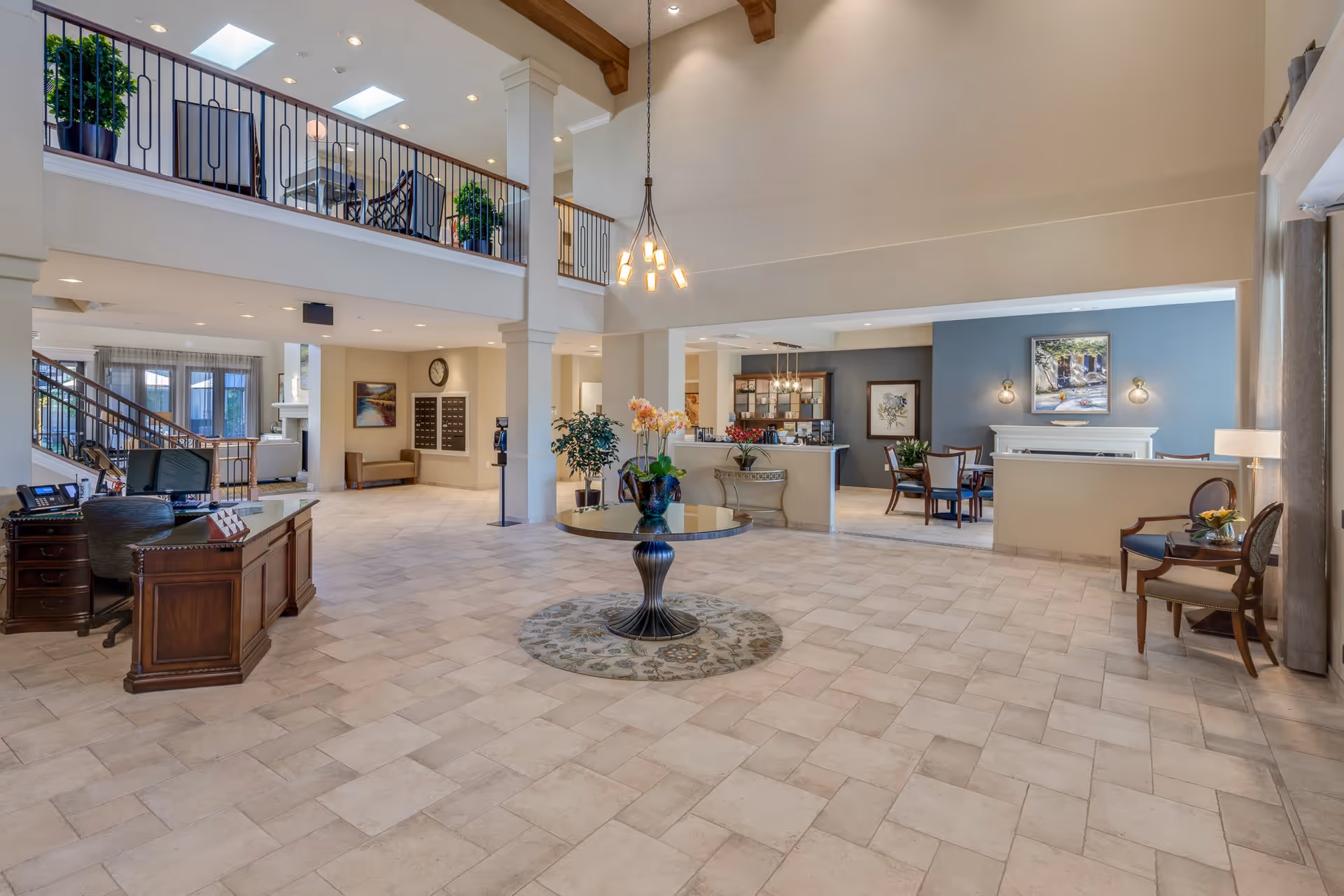 Spacious and well-lit senior living facility lobby with a central round table adorned with flowers, a wooden reception desk on the left, seating areas with chairs and tables on the right, and a second-floor balcony with plants and additional seating. The space features tiled flooring, high ceilings with exposed wooden beams, and modern lighting fixtures.