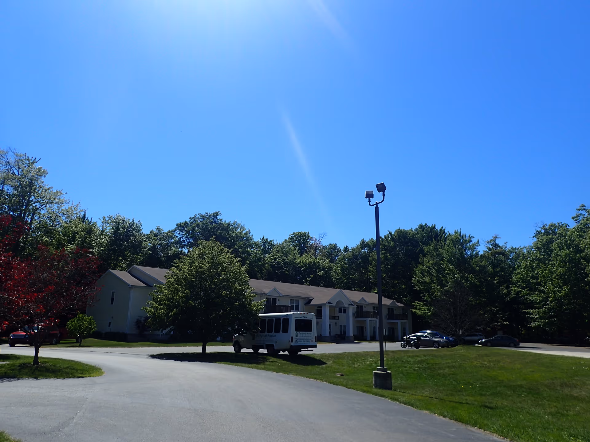 Two-story apartment building with a parking lot, a shuttle van, trees, and a clear blue sky.