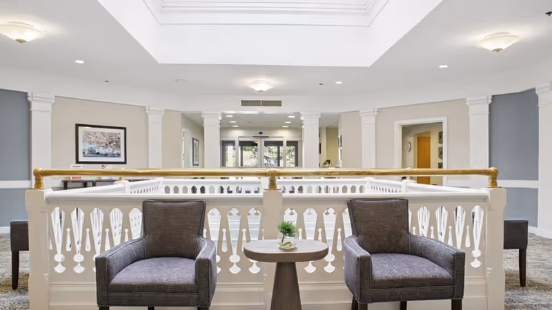 A bright and spacious interior common area in a senior living community featuring two gray upholstered armchairs with a small round wooden table between them. The area is surrounded by a white decorative railing with gold handrails. The background shows a hallway with doors and framed artwork on the walls, illuminated by ceiling lights.