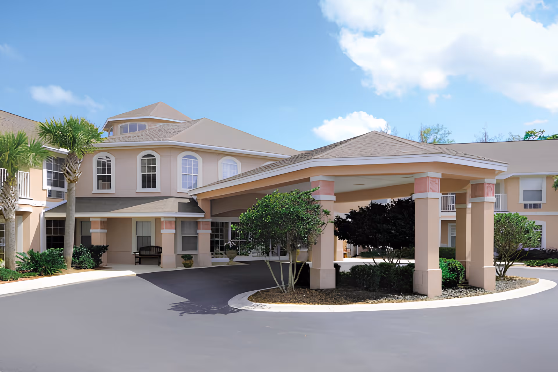Exterior view of Allegro Senior Living facility in Fleming Island, FL, showing a covered entrance with columns, landscaped bushes, palm trees, and a clear blue sky.