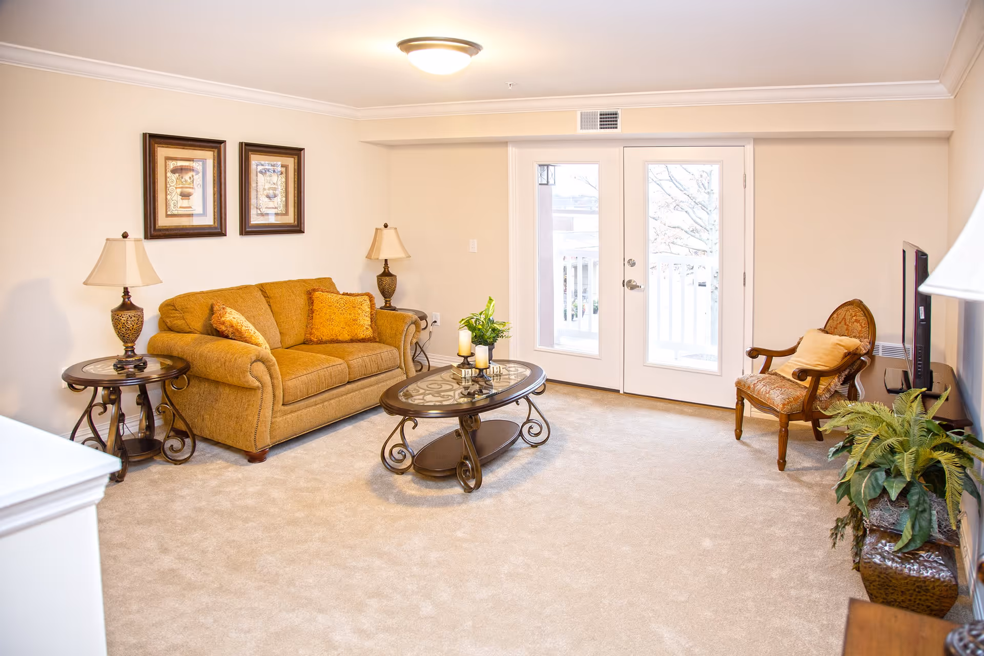 A cozy living room with beige walls and carpet, featuring a mustard-colored sofa with two decorative pillows, two framed pictures on the wall above the sofa, two matching table lamps on either side of the sofa, a glass-top coffee table with candles and a small plant, a patterned armchair with a yellow pillow, a flat-screen TV on a stand, and a large potted plant. French doors with glass panels lead to an outdoor balcony.